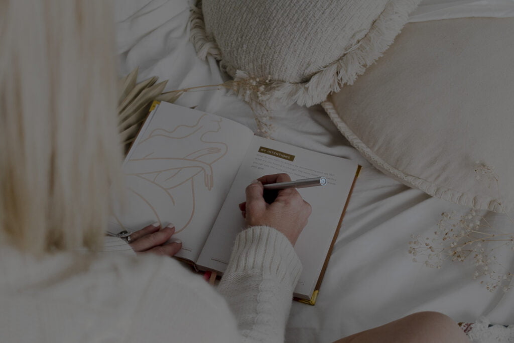blonde woman sitting on the bed writing in a book with her back to the camera.