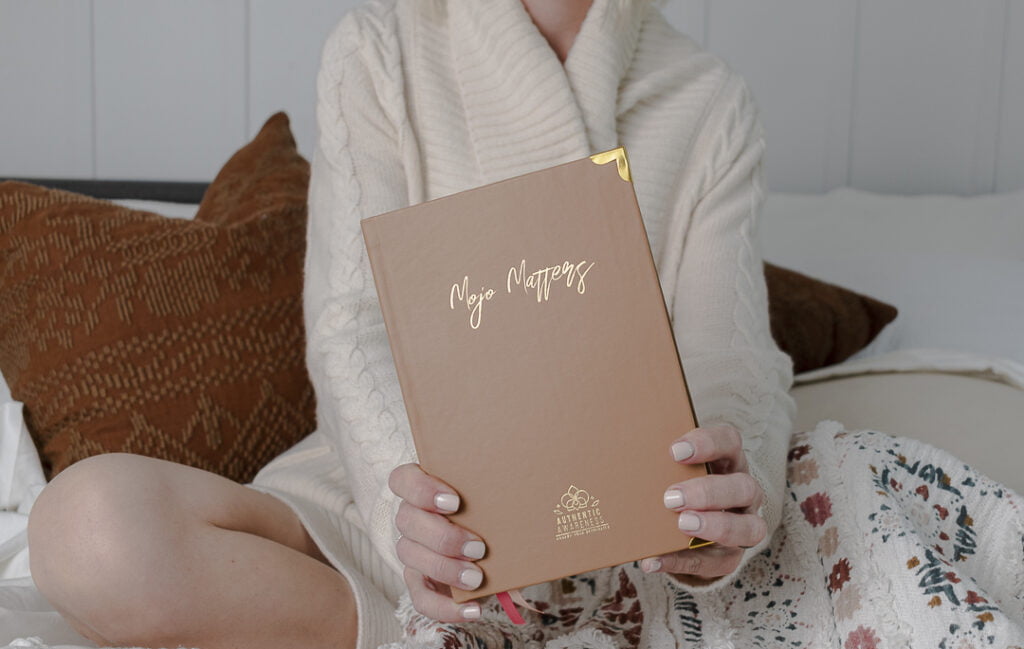 woman sitting cross legged holding a book out in front with the words Mojo Matters on the cover. She is holding the sex journal / sensuality journal.