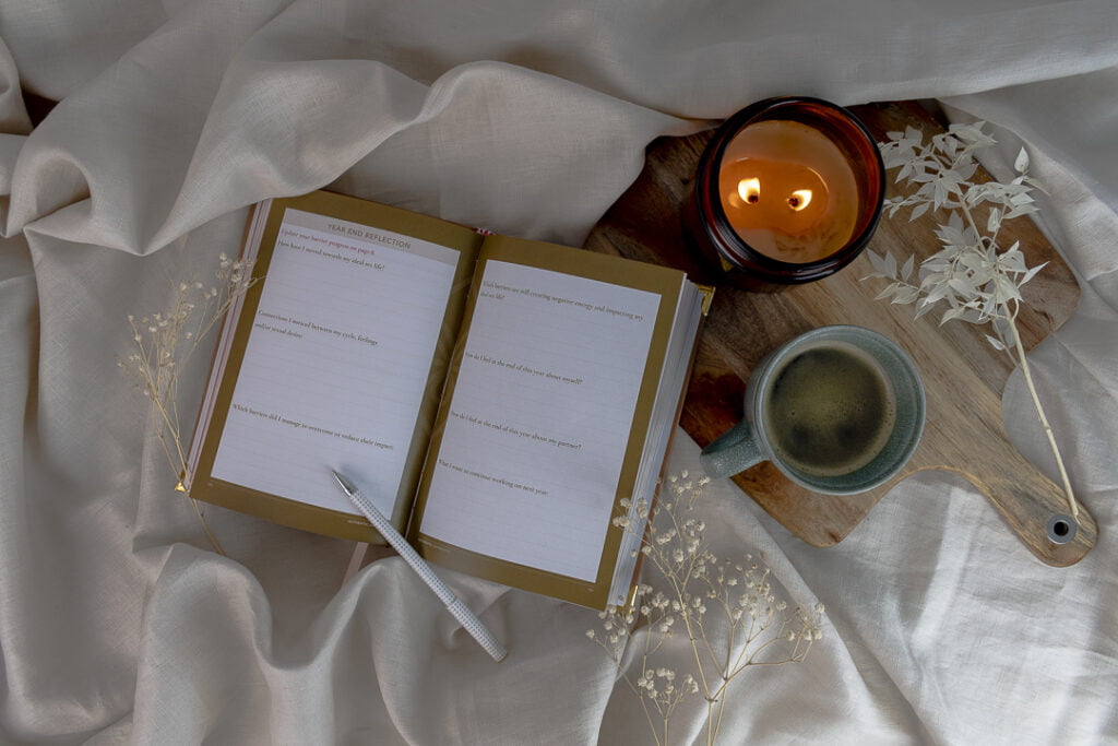 open book sitting on a sheet with a silver pen. candle burning and coffee next to book on a wooden board.