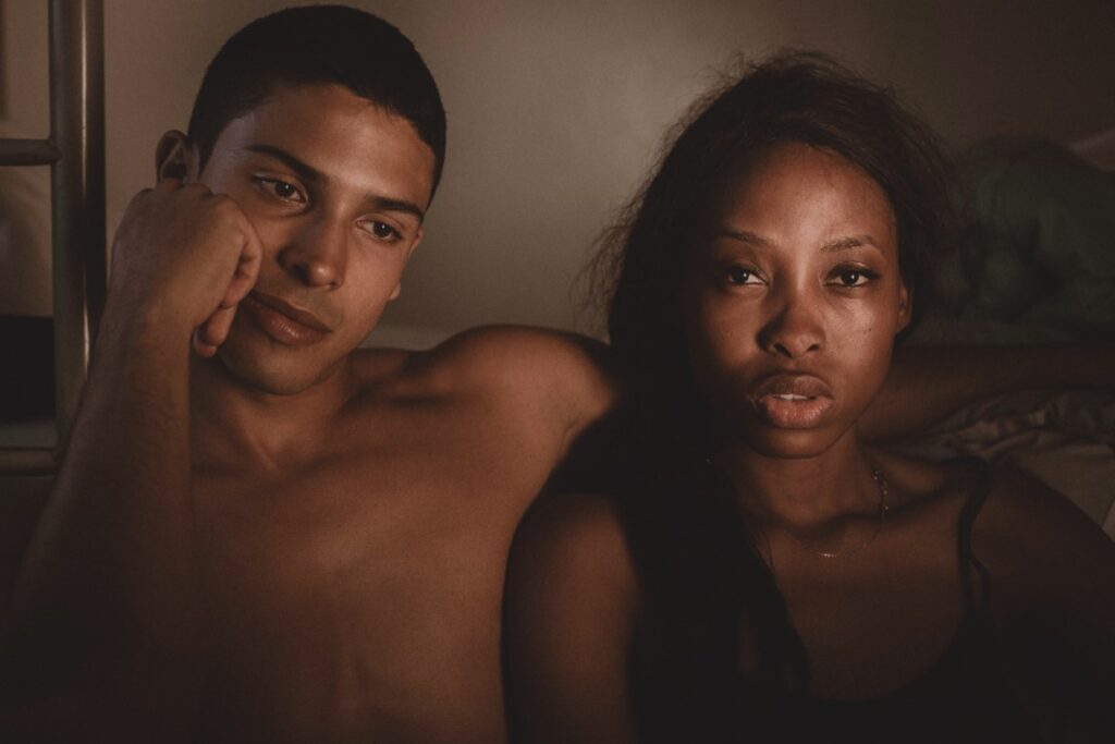 black woman and black man sitting next to each other. Woman is looking at the camera looking sad and mad is resting his head on his fist looking down away from the camera. Woman have issues with sexual desire