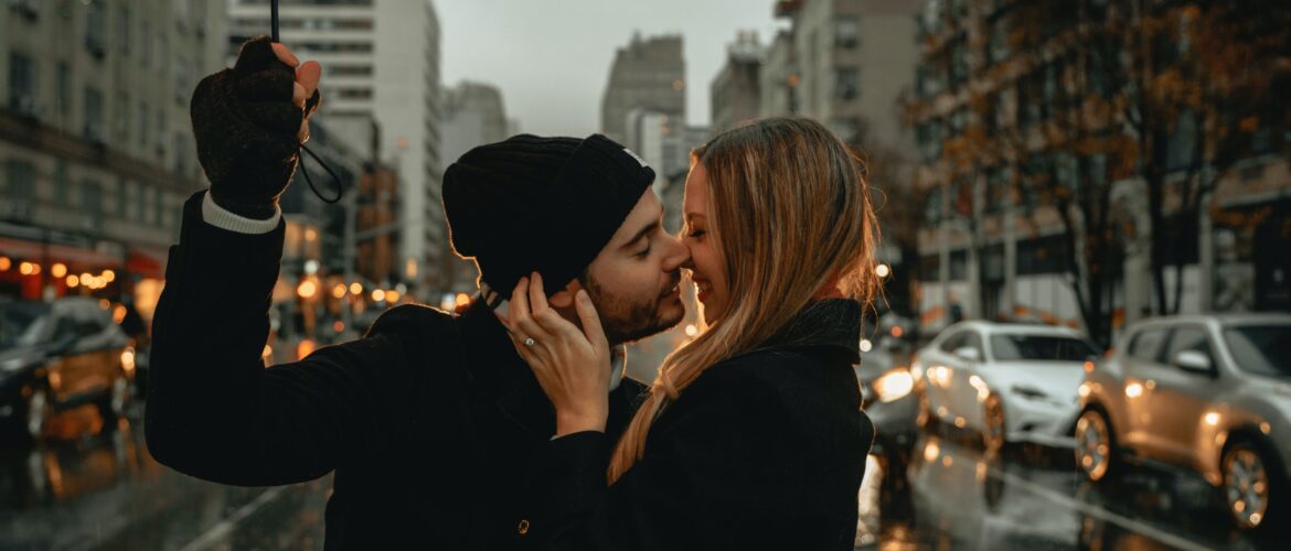 man and woman standing in the middle of a street boost intimacy by kissing. The man is holding an umbrella and there are cars and buildings behind the couple.