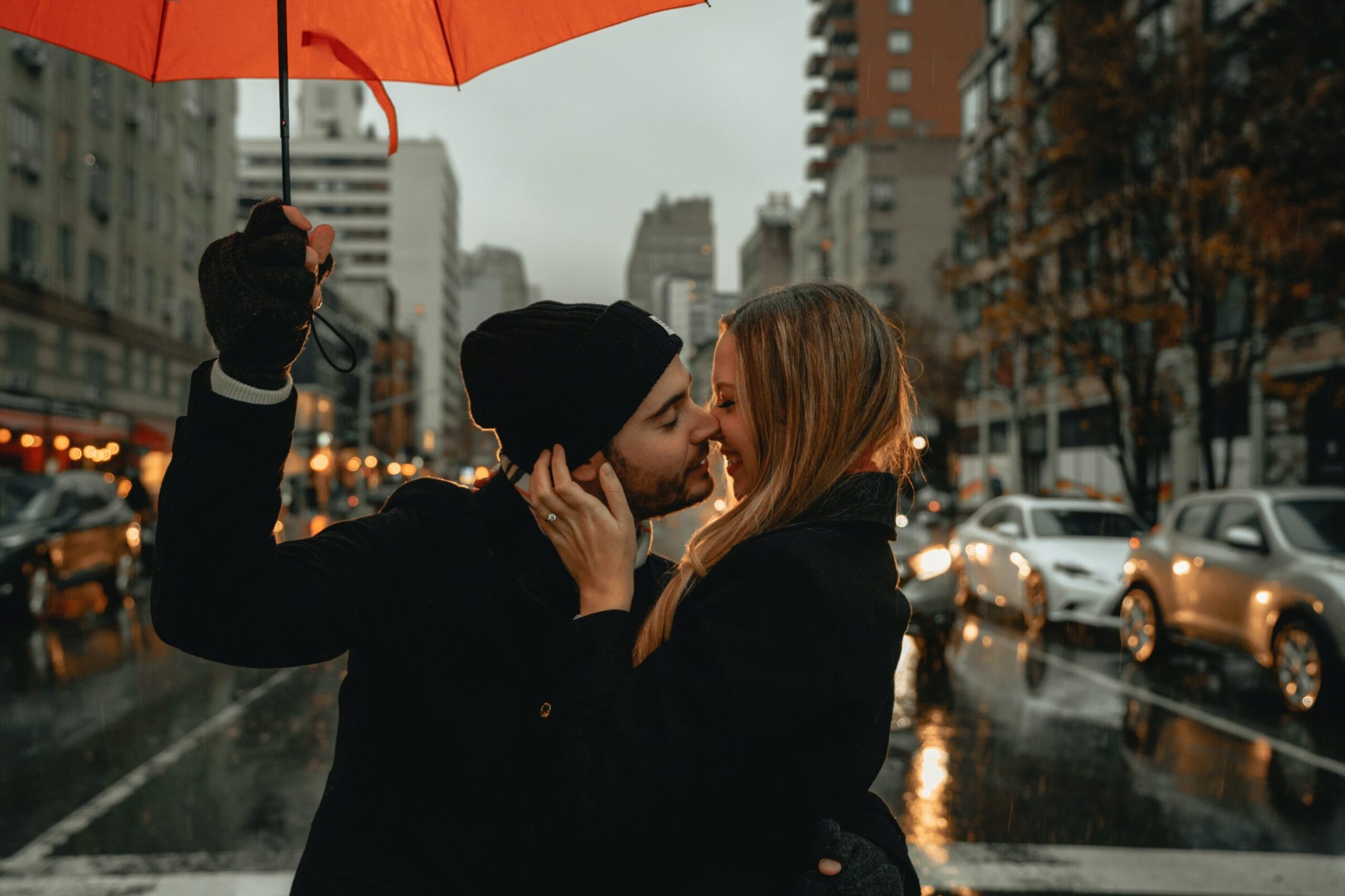 man and woman standing in the middle of a street boost intimacy by kissing. The man is holding an umbrella and there are cars and buildings behind the couple.