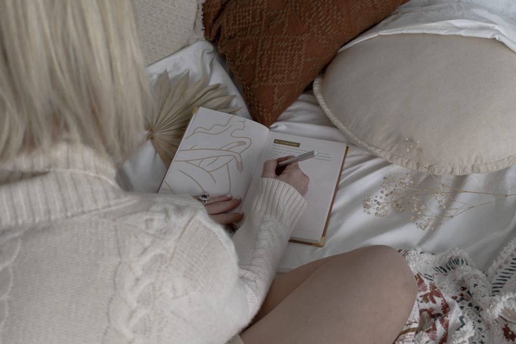 blonde woman sitting on a bed writing in a journal with her back to the camera. writing about her menstrual cycle and sex life.