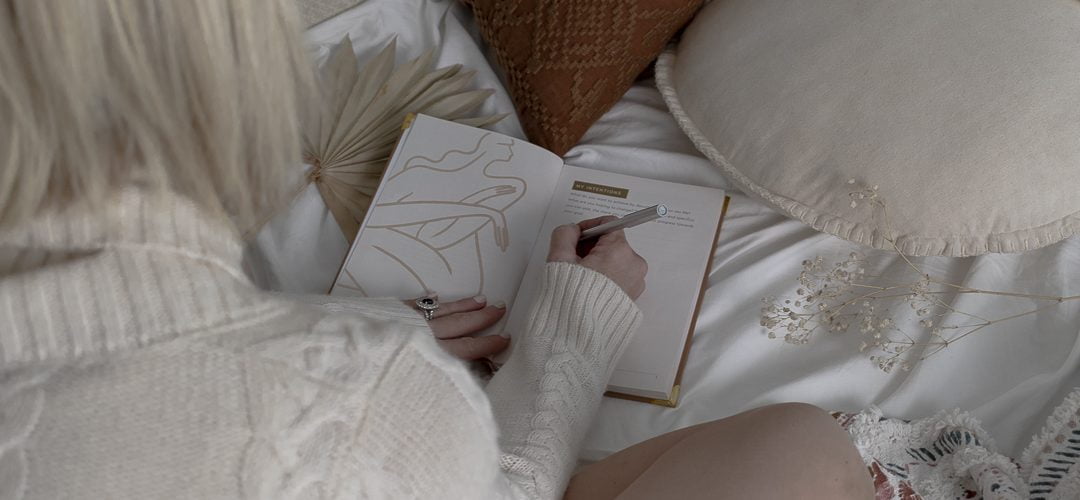 blonde woman sitting on a bed writing in a journal with her back to the camera. writing about her menstrual cycle and sex life.