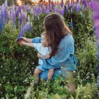 woman sitting in a field of flowers holding a toddler. woman and toddler are touching a purple flower