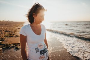 pregnant woman standing on the beach looking at the water with sunglasses on her head wearing a t-shirt saying I'm Coming