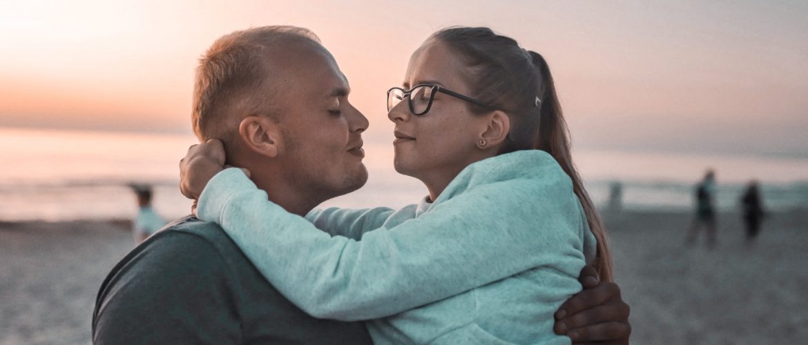 man carrying his female sex partner on the beach. The woman is wearing glasses with her arms around the mans neck whilst the man and woman look at each other