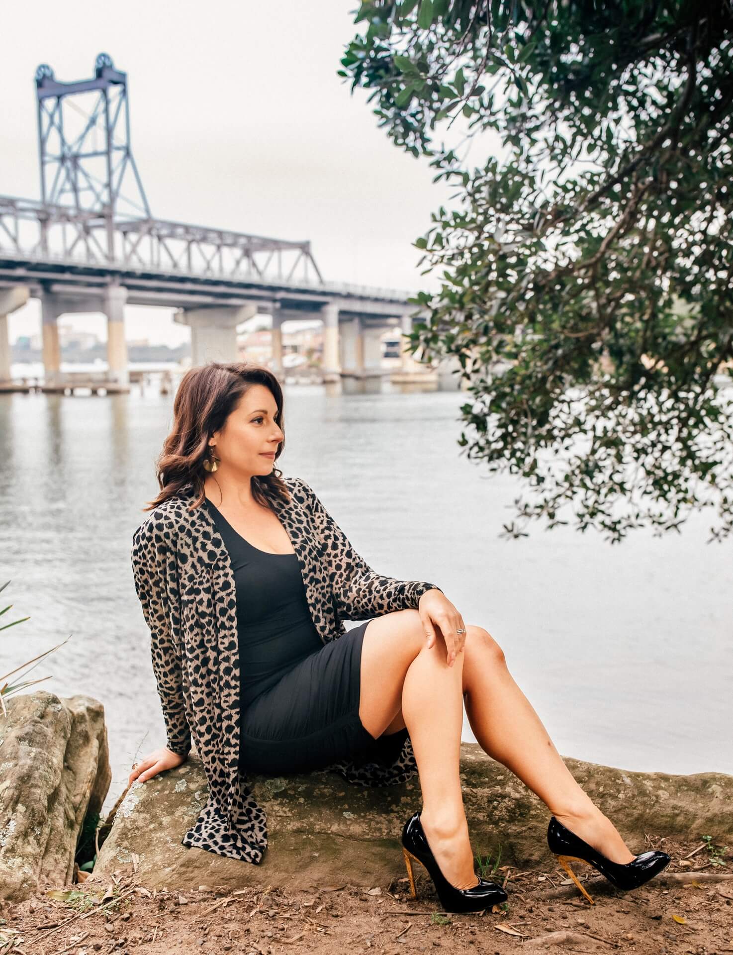 sexually confident woman wearing heels sitting on a rock looking to the side in front of water with a bridge in background