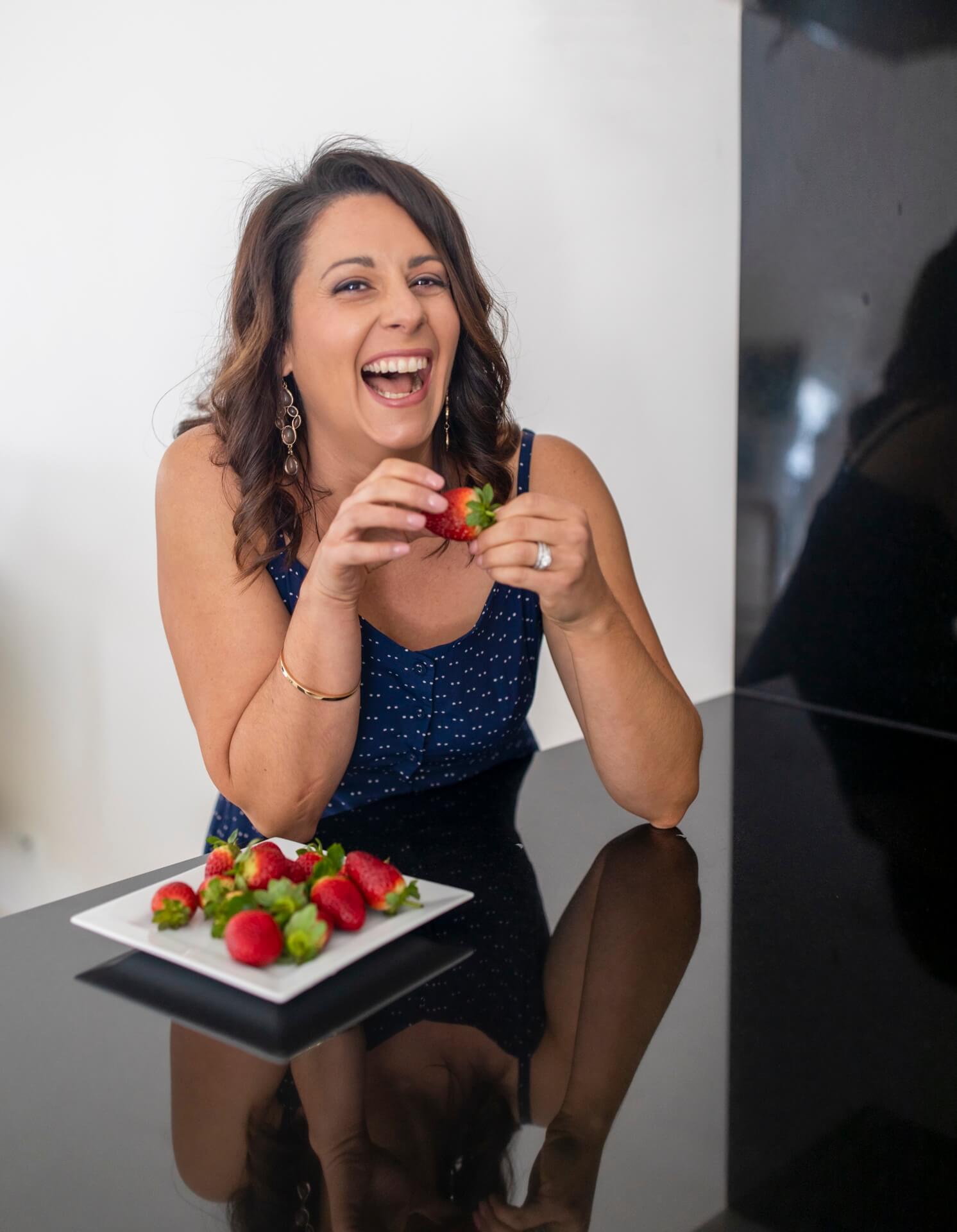 woman sitting at the bench laughing with a plate of strawberries and a strawberry in hand