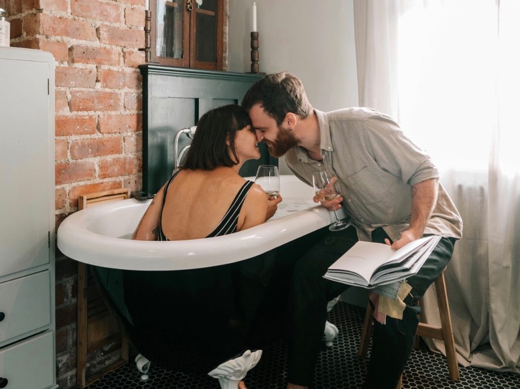 woman sitting in bathtub with her back to camera. man sitting on a stool next to bath tub with an open book. woman and man with glass of wine in their hand leaning towards each other to kiss