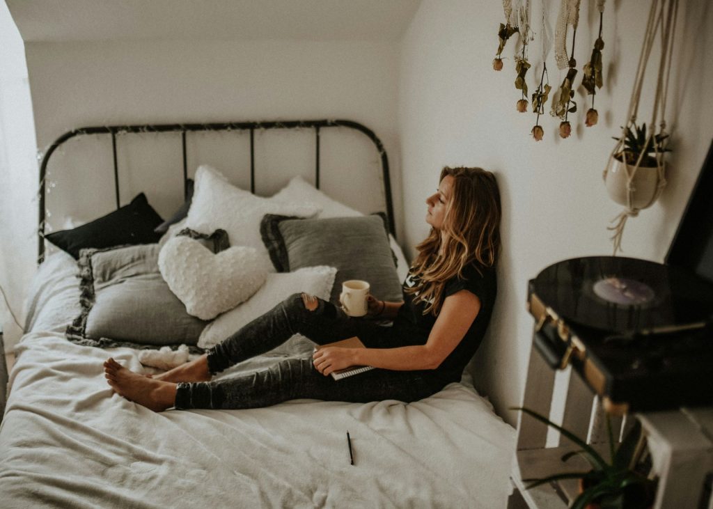 woman sitting on bed with her back against a wall. Legs stretched out in front holding a coffee on one knee. Woman staring across the room.