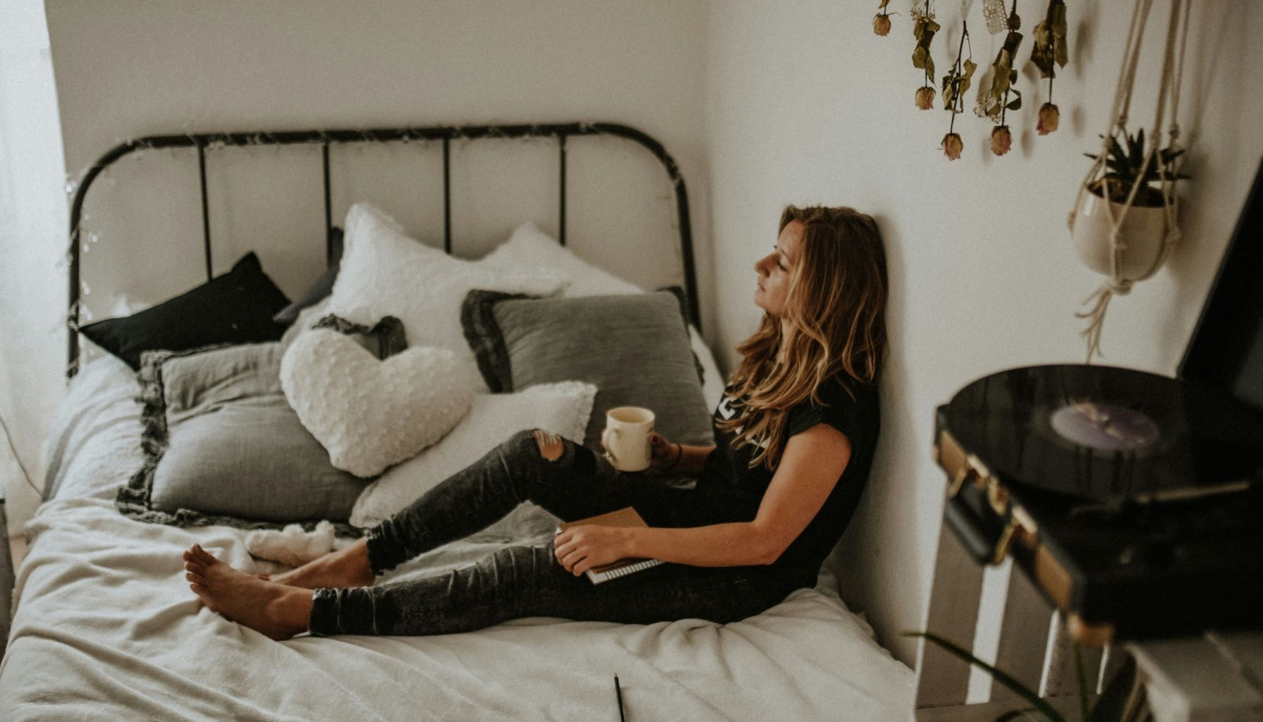 woman sitting on bed with her back against a wall. Legs stretched out in front holding a coffee on one knee. Woman staring across the room.