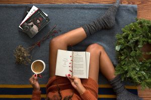 book resting on a woman's legs. Woman is sitting on the floor with a cup of coffee and more books to the side