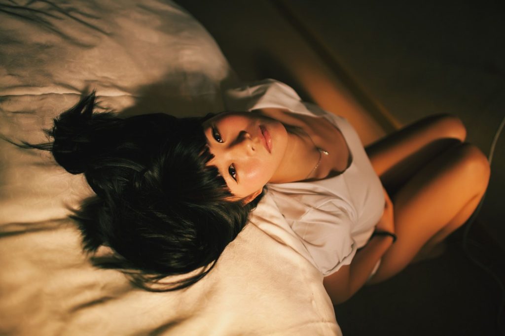 asian woman sitting on the floor, leaning on the bed looking up to the camera. Woman wearing a white t-shirt with bent knees. Woman looking at camera leaning on bed looking to boost libido