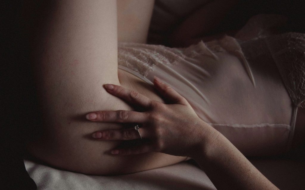 woman lying on her back in lingerie on the bed with her hand on her hip exploring self pleasure and her body. Side view of woman in sheer lingerie.