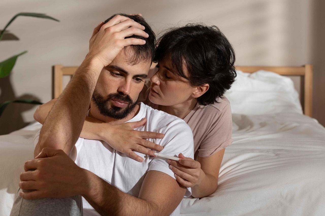 man and woman on bed looking sad. man sitting in front of woman. Woman has her arms around man hugging him from behind.