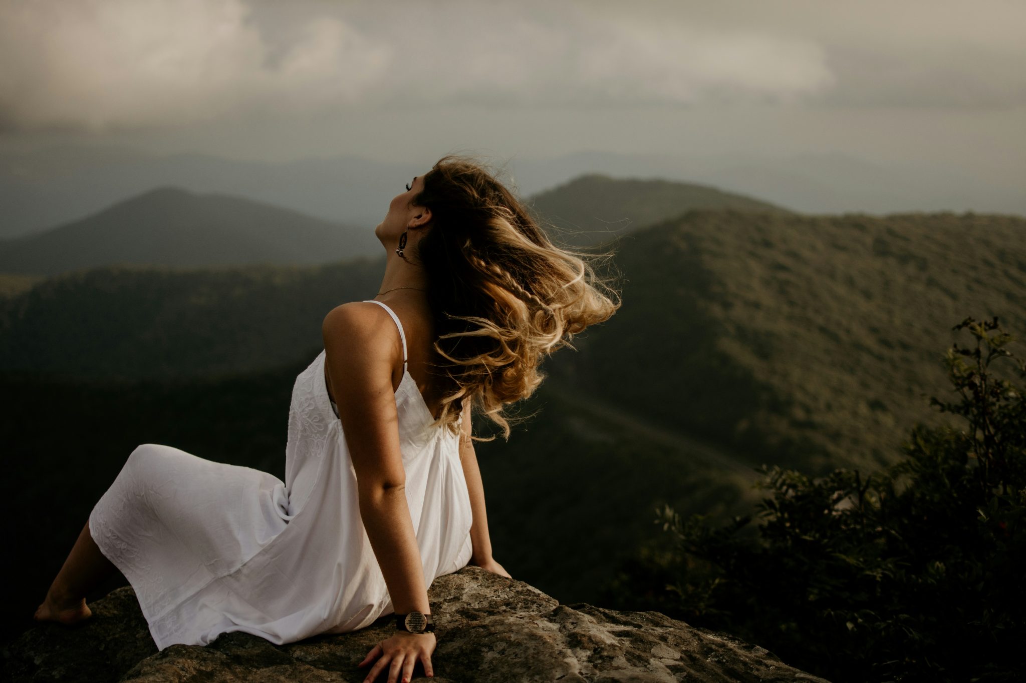 woman sitting on top of a mountain in a white dress with her hair flowing in the wind. She is leaning back with her back to the camera