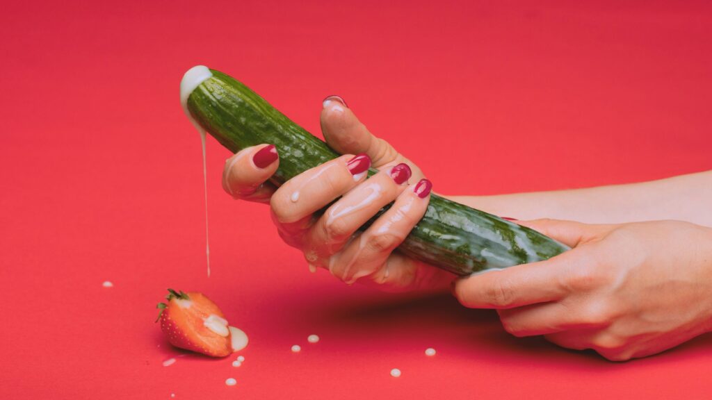 female hands holding a culcumber with a condom on it. a cut strawberry with white drops sits underneath the cucumber head which is leaking a white substance representing ejaculation from a penis