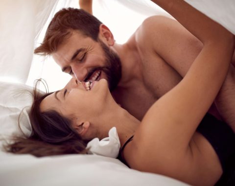unleash her ecstasy - mastering the art of female pleasure. woman lying on her back in bed, man next to her. Man and woman are laughing while male gazes down at woman. Man and woman under sheet